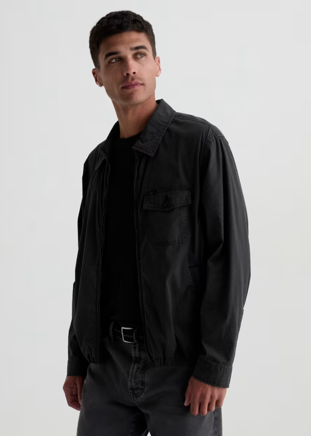 A man wearing a black utility-style jacket over a black shirt and dark gray jeans, photographed against a plain light gray studio background.