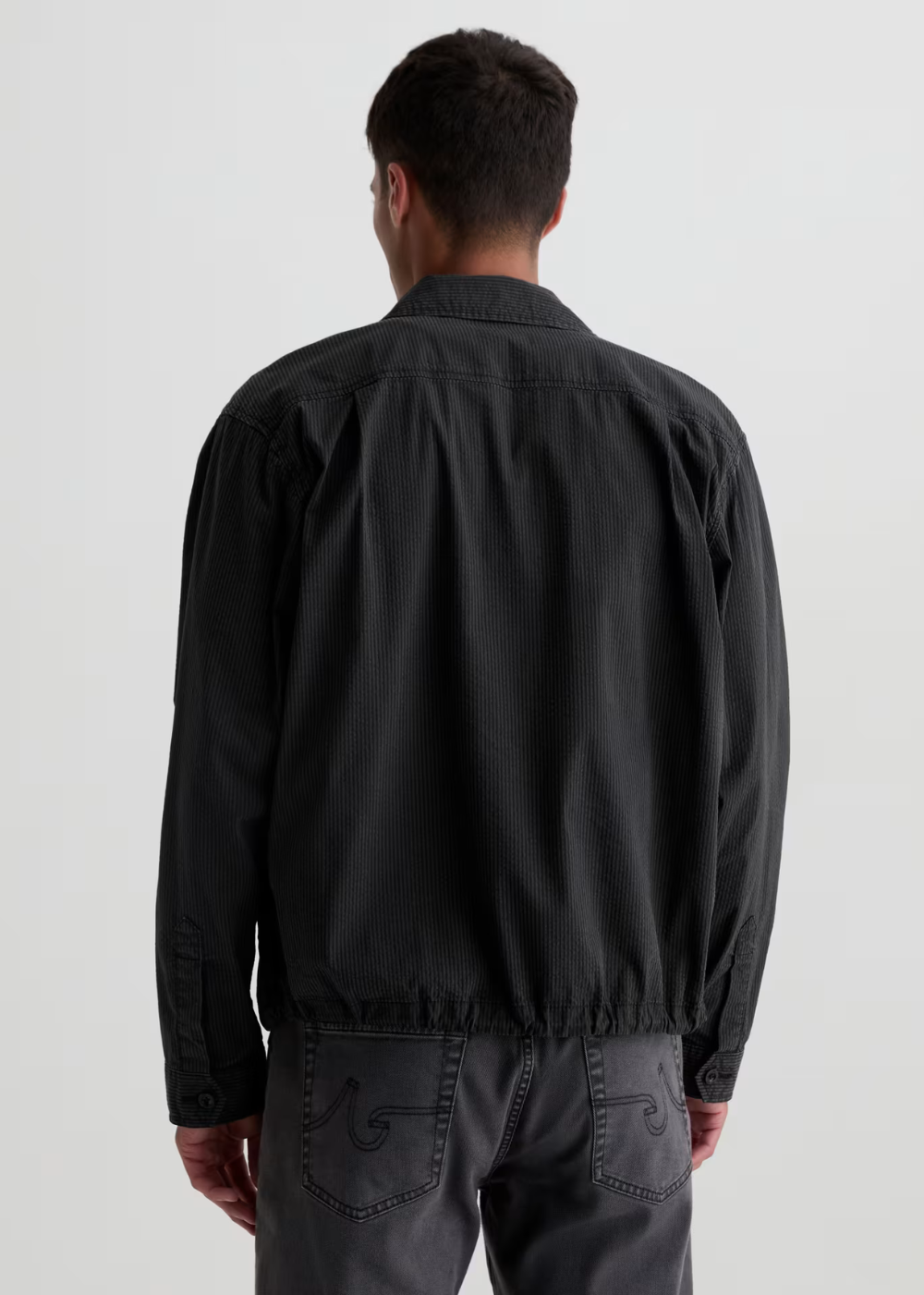 A man viewed from behind wearing a black lightweight jacket and dark gray trousers against a plain light gray studio background.