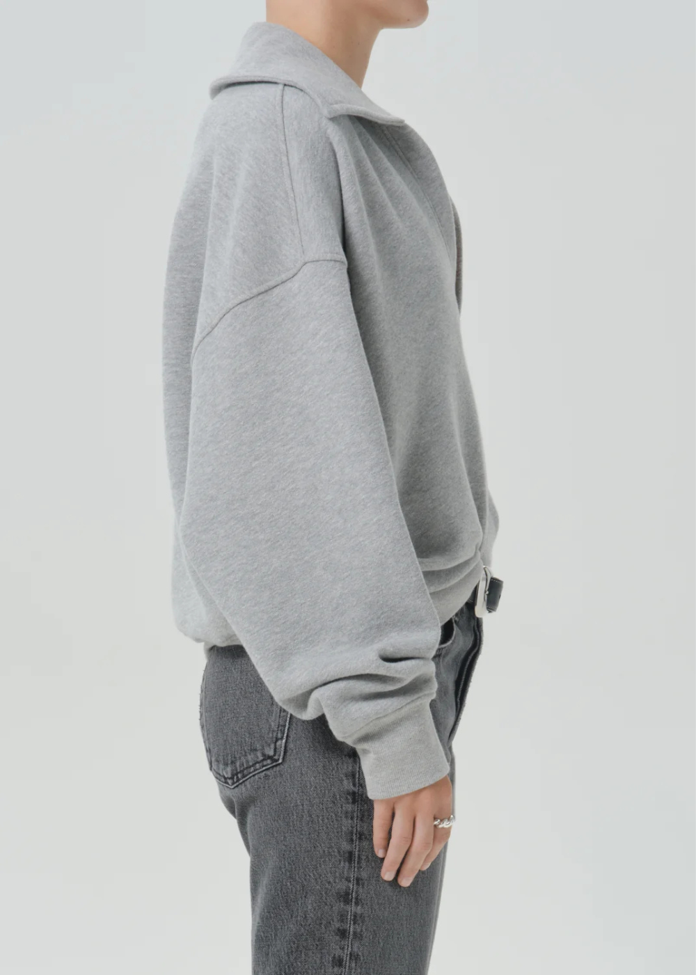 A side-profile view of a person wearing a light gray oversized hoodie and dark gray jeans against a plain light gray studio background.