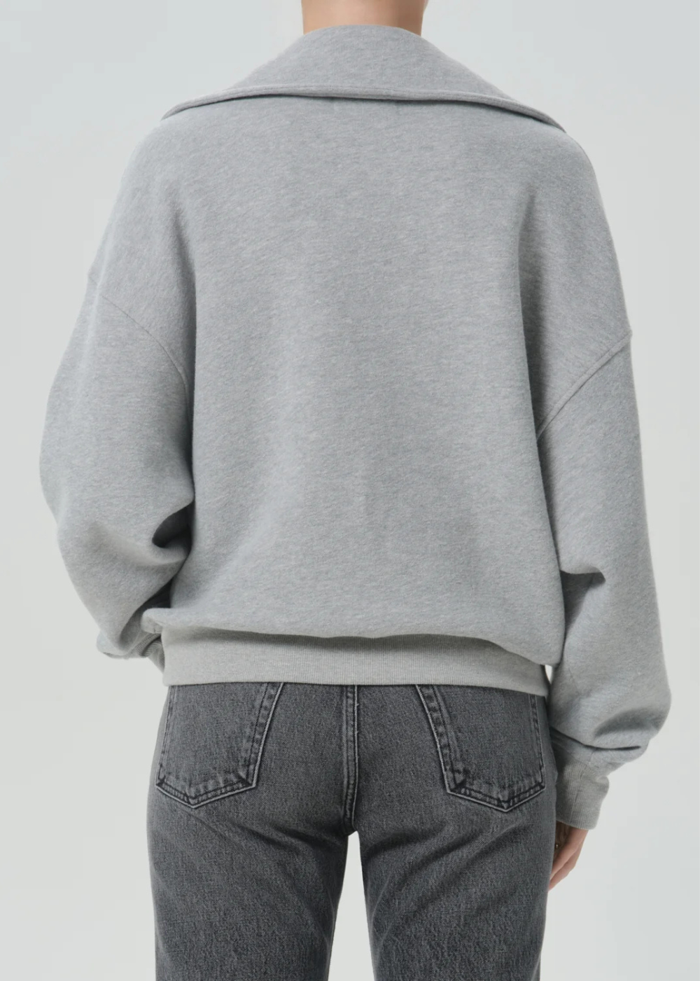 A person shown from the back wearing a light gray oversized knit top or sweatshirt with a wide collar and dark gray jeans, against a plain light gray studio background.