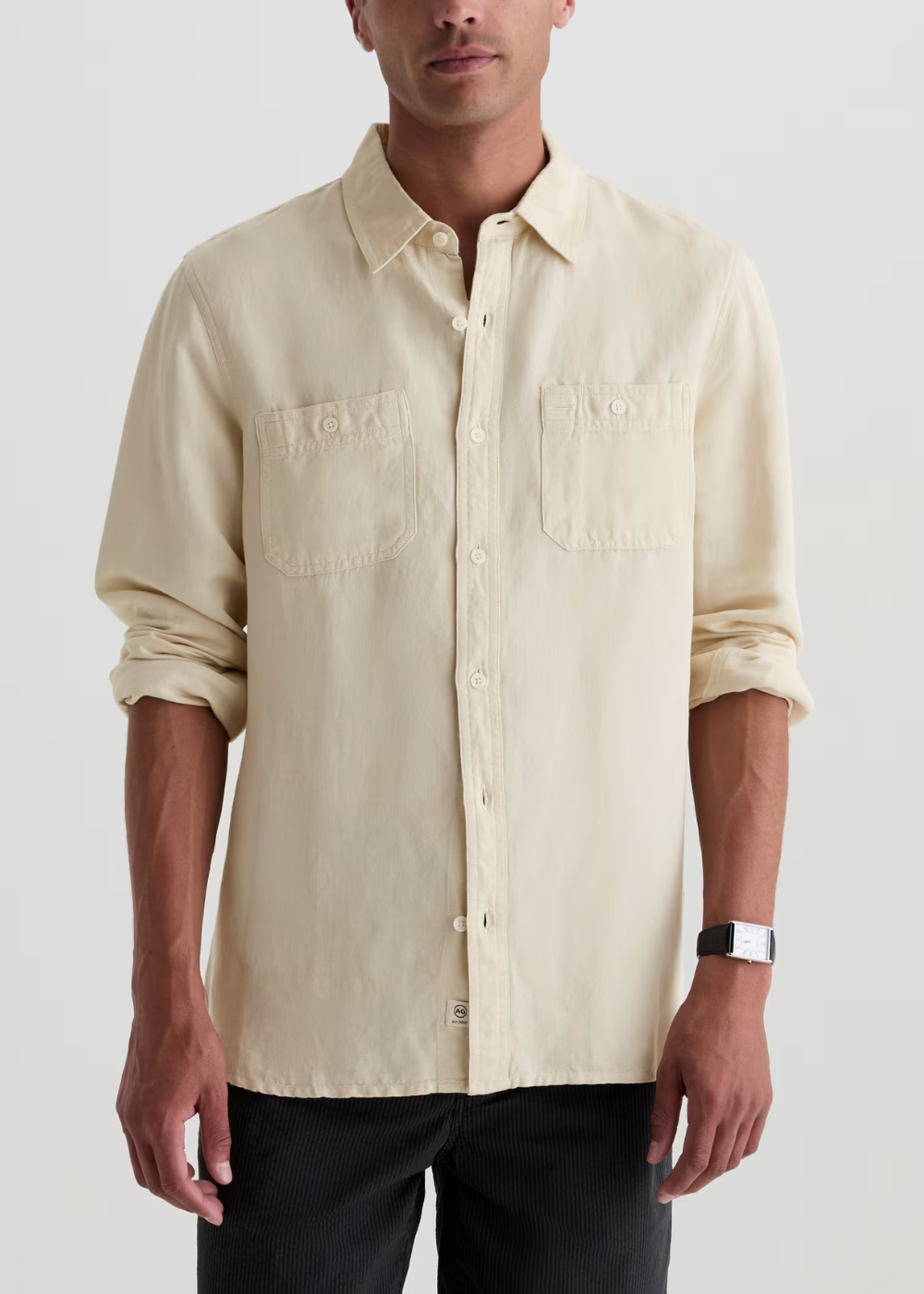 A man wearing a light cream-colored, long-sleeve linen button-up shirt with two chest pockets, paired with black pants, against a plain light gray background.