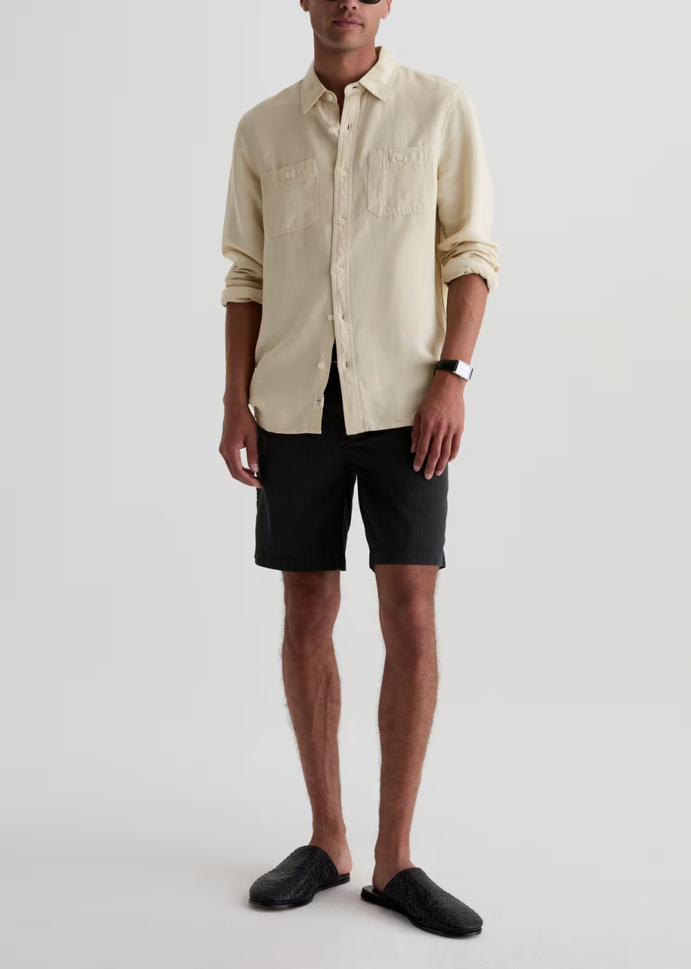 A man modeling a light beige long-sleeve button-up shirt with black shorts and black slip-on shoes, standing against a plain light gray studio background.