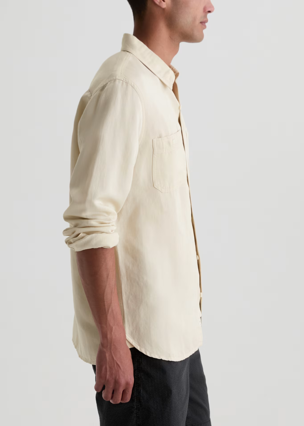 A man shown in side profile wearing a light cream-colored linen button-down shirt with rolled-up sleeves and dark trousers, set against a plain light gray studio background.