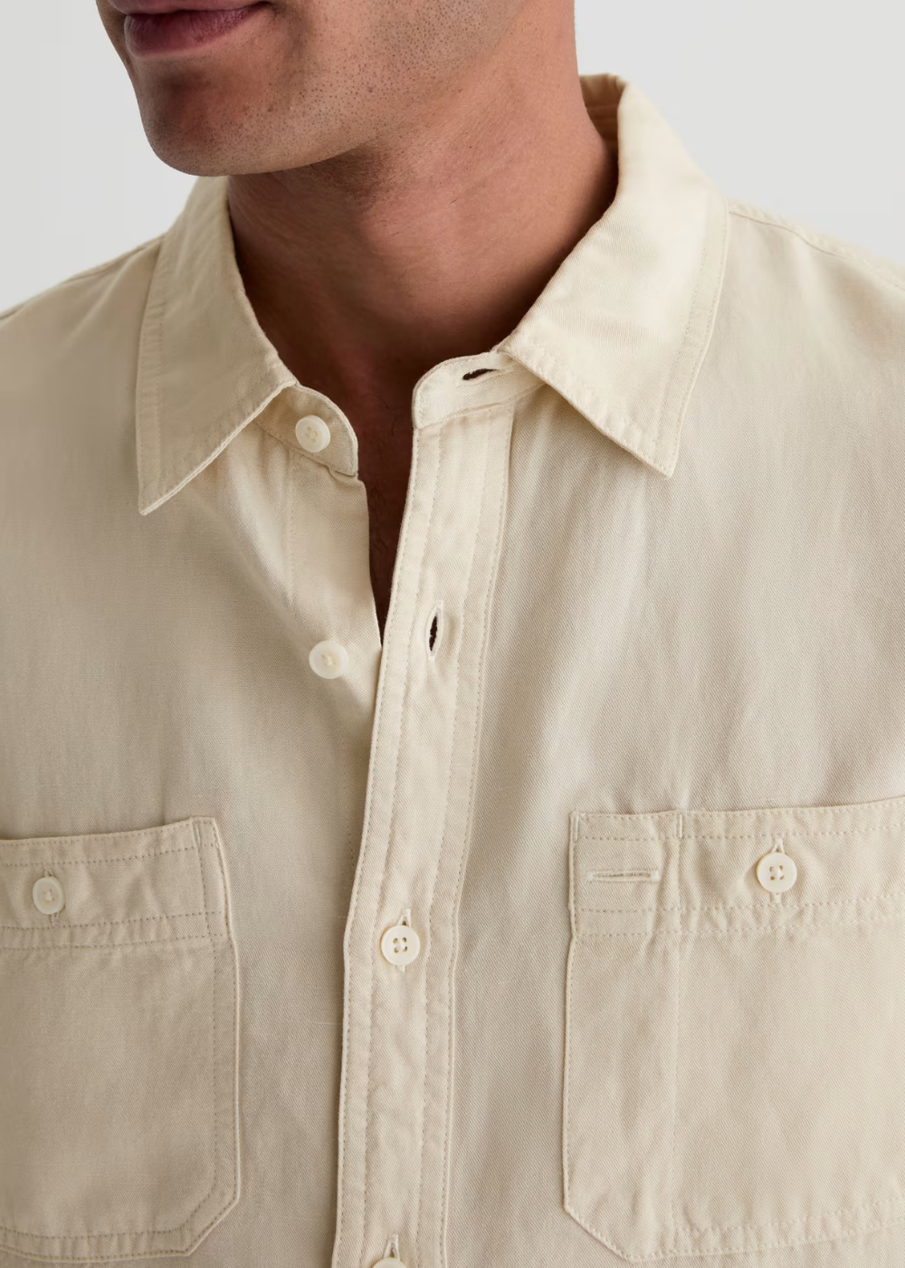 Close-up of a man wearing a light beige or cream-colored button-down shirt with two chest pockets and white buttons, against a plain light gray background.