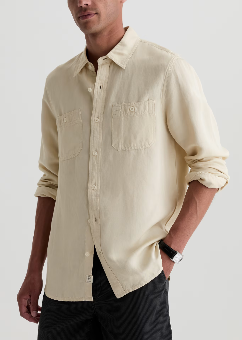 A man wearing a light cream-colored long-sleeve linen shirt with two buttoned chest pockets, paired with dark trousers, against a light gray studio background.