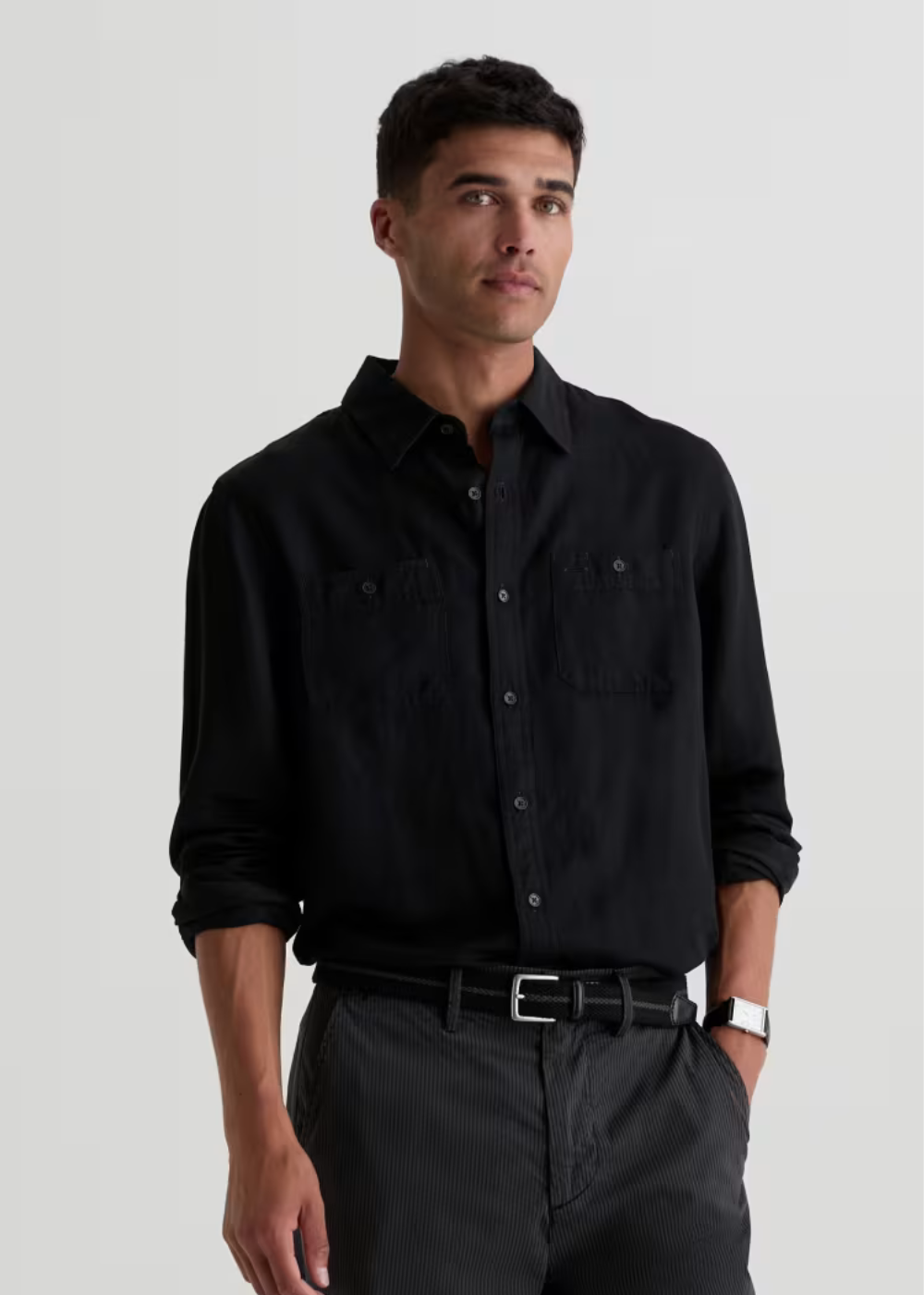 A man wearing a black long-sleeve button-down shirt with chest pockets, paired with dark gray trousers and a black belt, photographed against a light gray studio background.