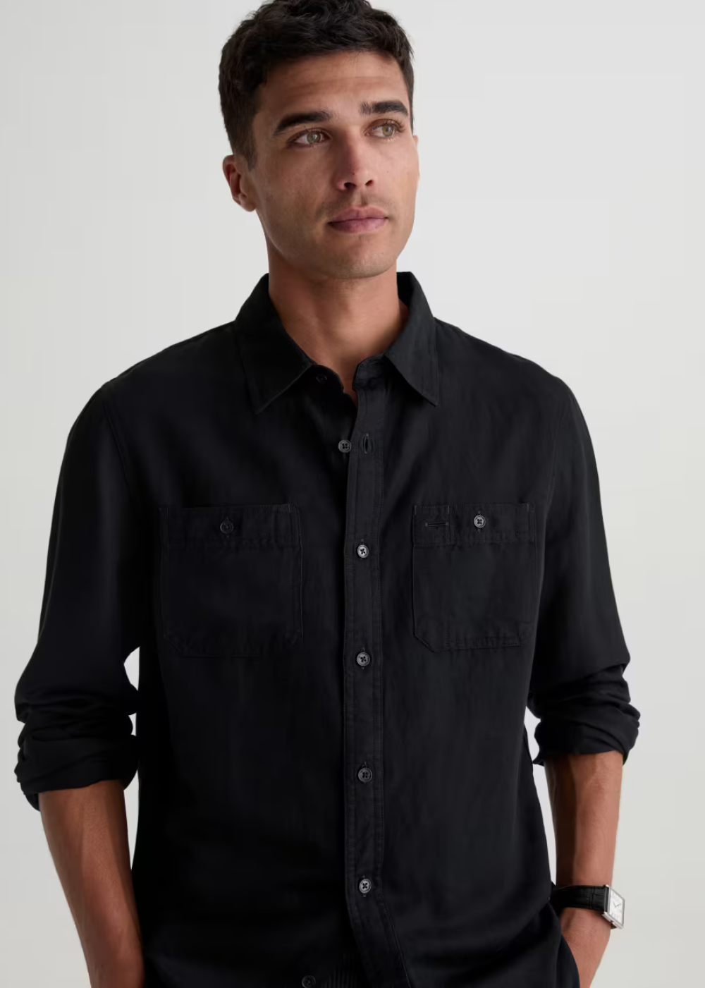 A man wearing a black long-sleeve button-up shirt with two chest pockets, photographed against a plain light gray studio background.