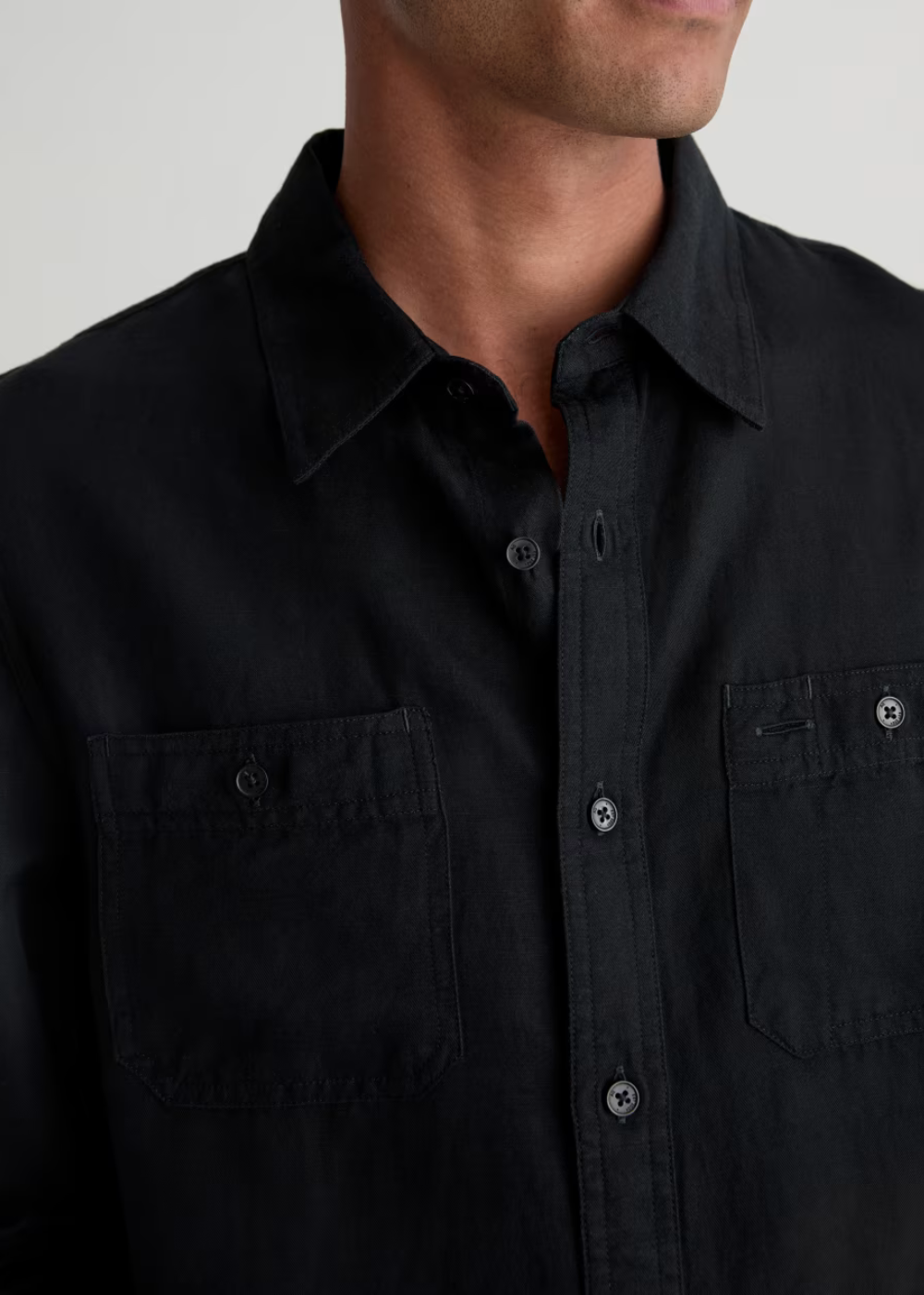Close-up of a man wearing a black button-up shirt with two chest pockets and dark buttons, against a light gray background.