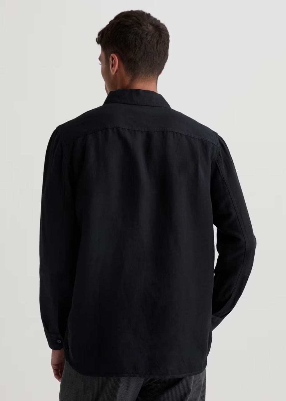 A man viewed from the back wearing a black long-sleeve button-up shirt and dark trousers, photographed against a plain light gray studio background.