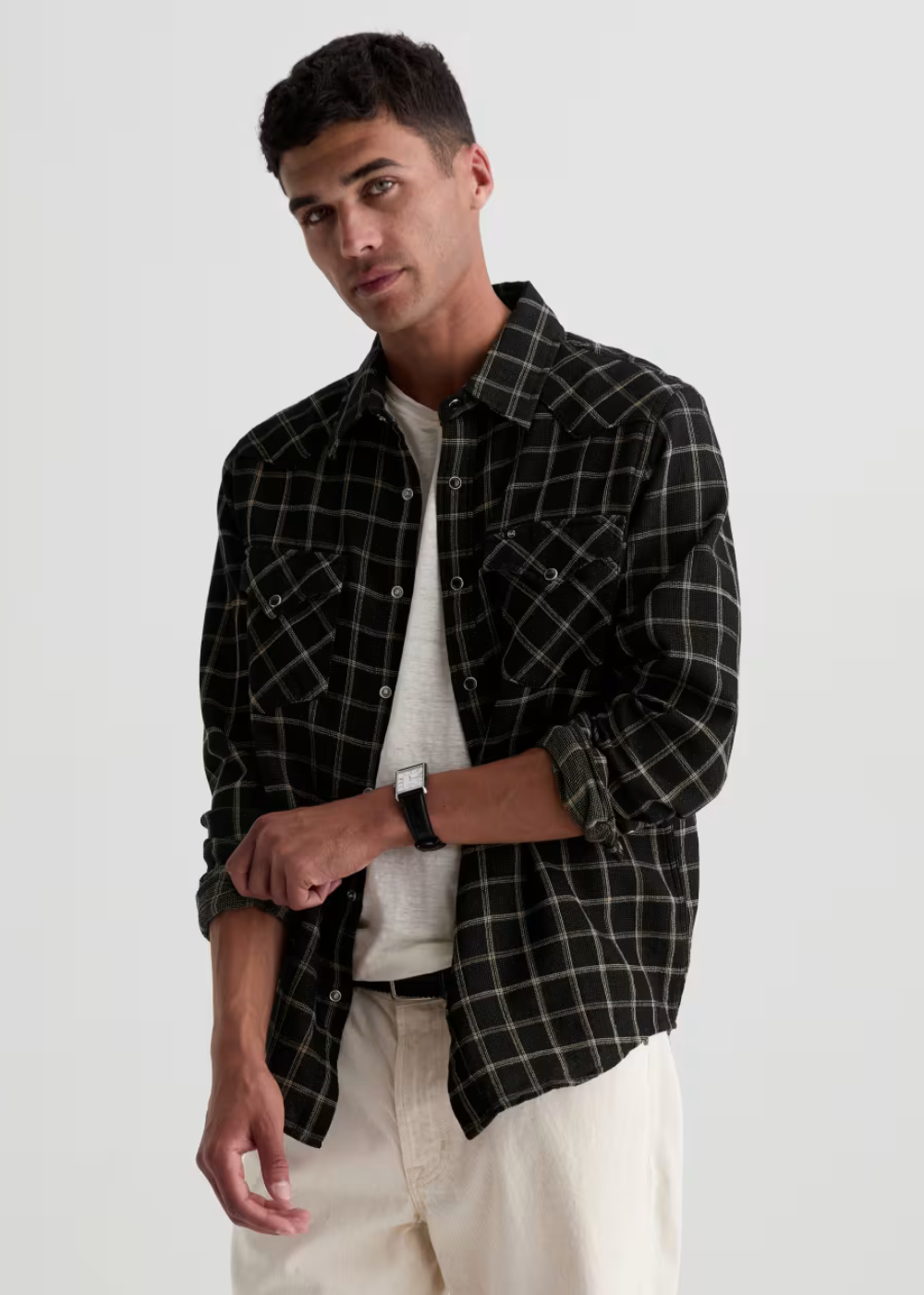 A man wearing a black and white plaid button-down shirt over a white t-shirt and cream-colored pants, photographed against a light gray studio background.
