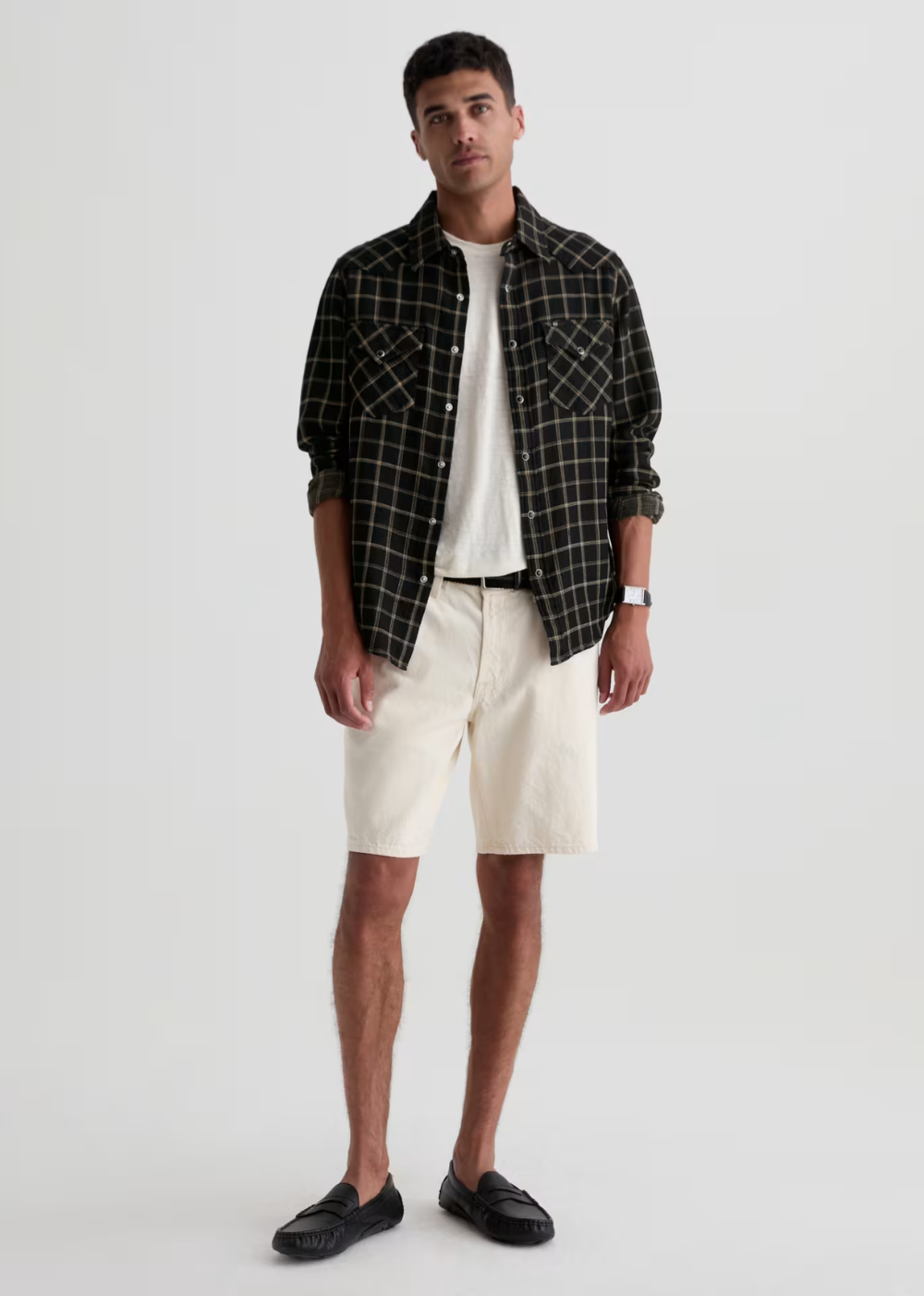 A man modeling a black and white plaid button-down shirt over a white t-shirt, paired with cream-colored shorts and black loafers, against a plain light gray studio background.