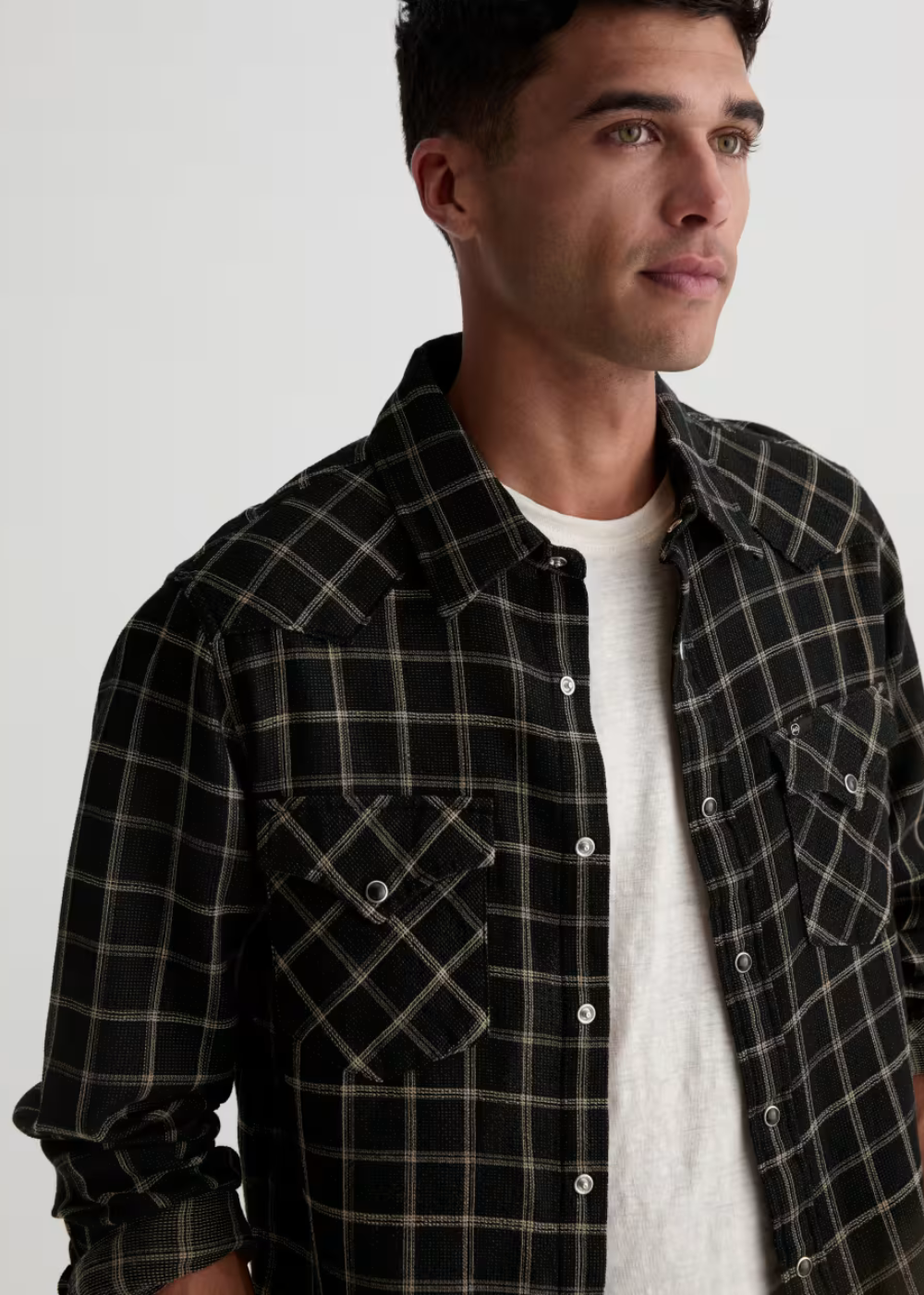 A man wearing a black and beige plaid button-down shirt over a white t-shirt, photographed against a light gray studio background.