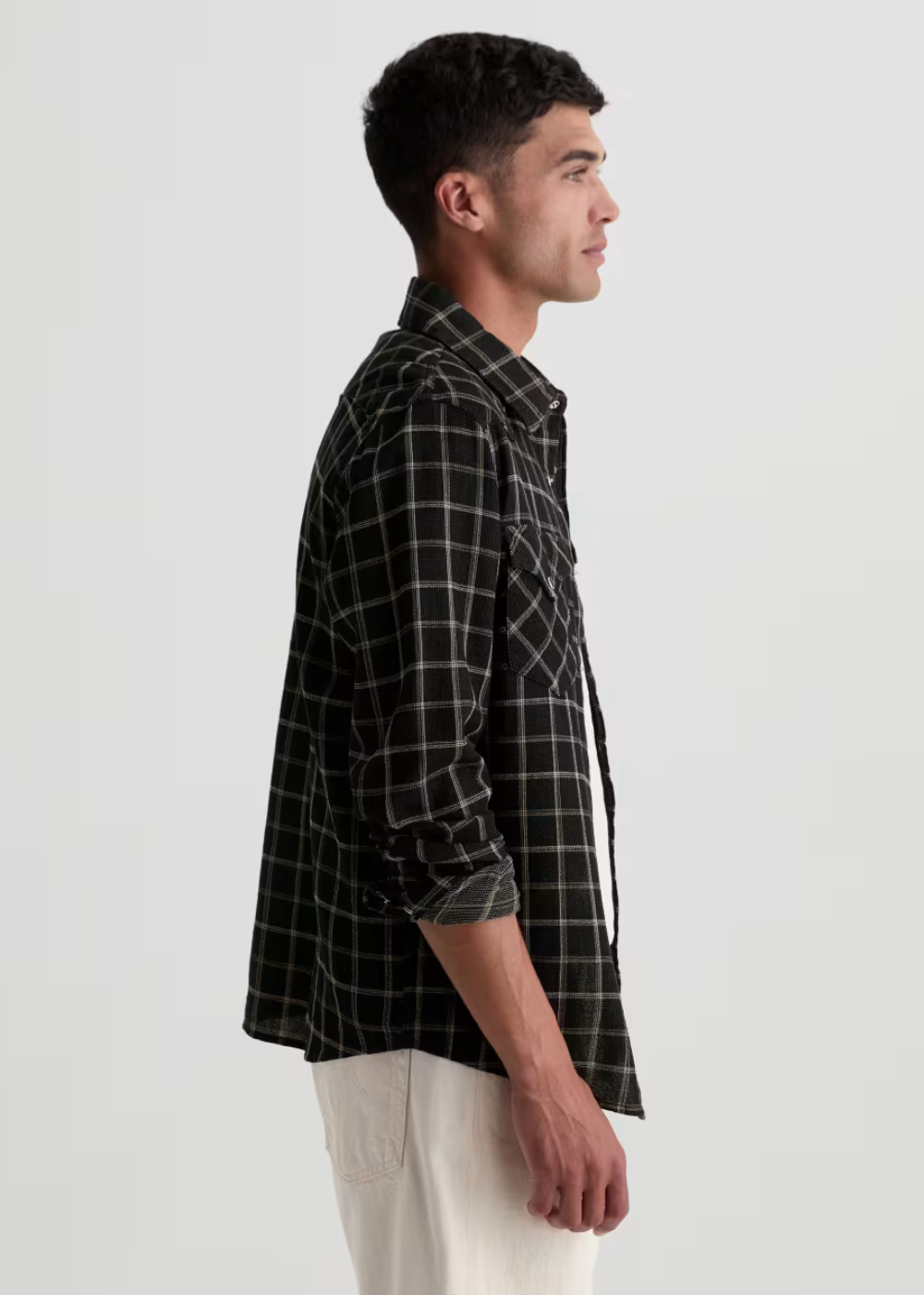 A man shown in profile wearing a black and white plaid button-down shirt with white pants, photographed against a plain light gray studio background.