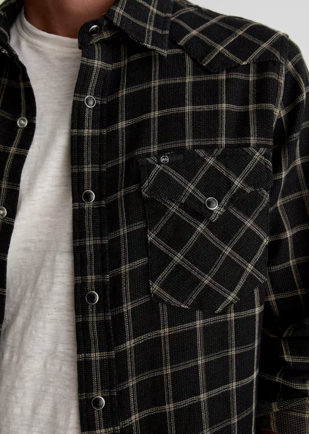 A close-up of a person wearing a black plaid button-up shirt over a light gray t-shirt. The shirt features a classic plaid pattern with thin beige lines and silver metal buttons.