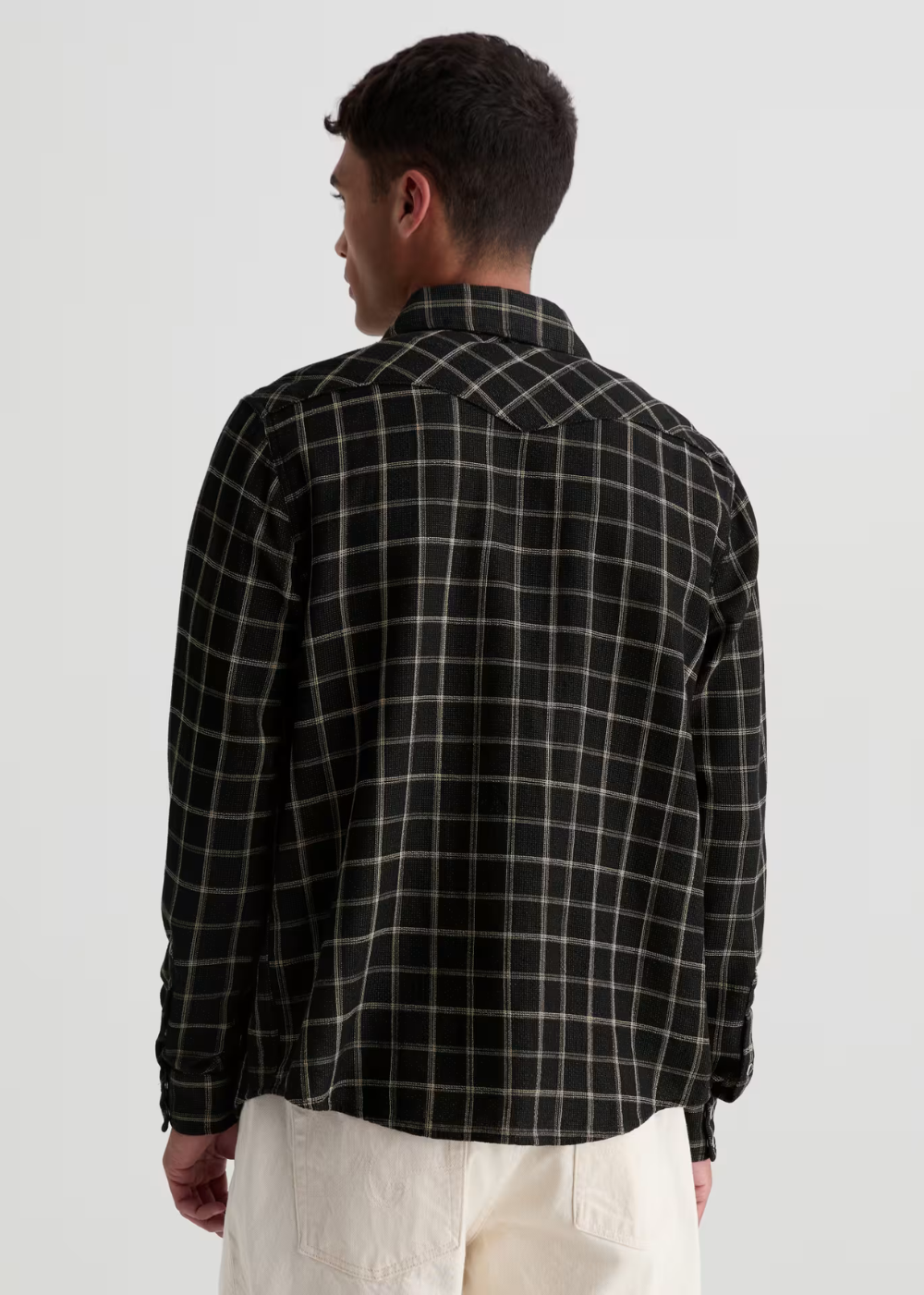 A man viewed from the back wearing a black long-sleeve plaid shirt with thin white grid lines and off-white trousers, against a plain light gray studio background.