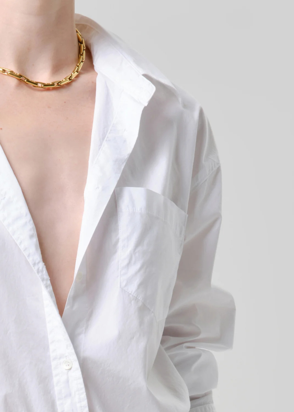 A close-up of a person wearing a chunky gold chain necklace over a white button-down shirt, shown against a light gray background.