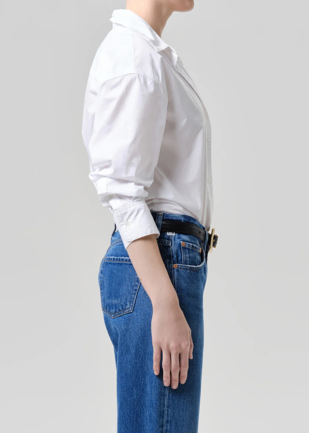 A side-profile view of a person wearing a white long-sleeve button-up shirt tucked into blue jeans with a black belt, against a plain light gray studio background.