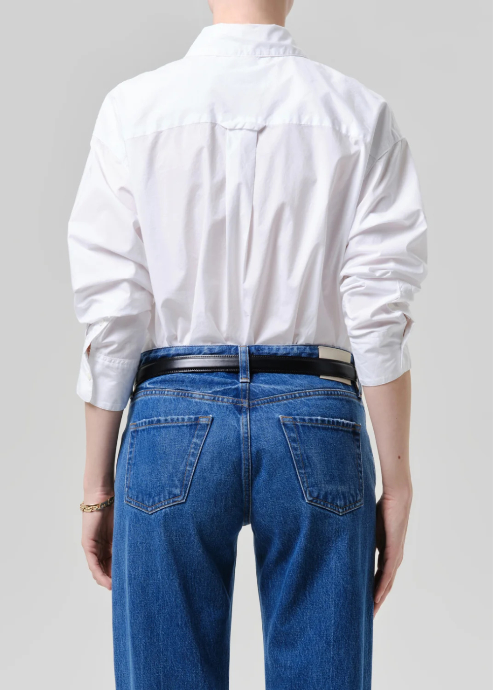 Back view of a person wearing a white long-sleeve button-up shirt tucked into blue jeans with a black belt, against a light gray studio background.