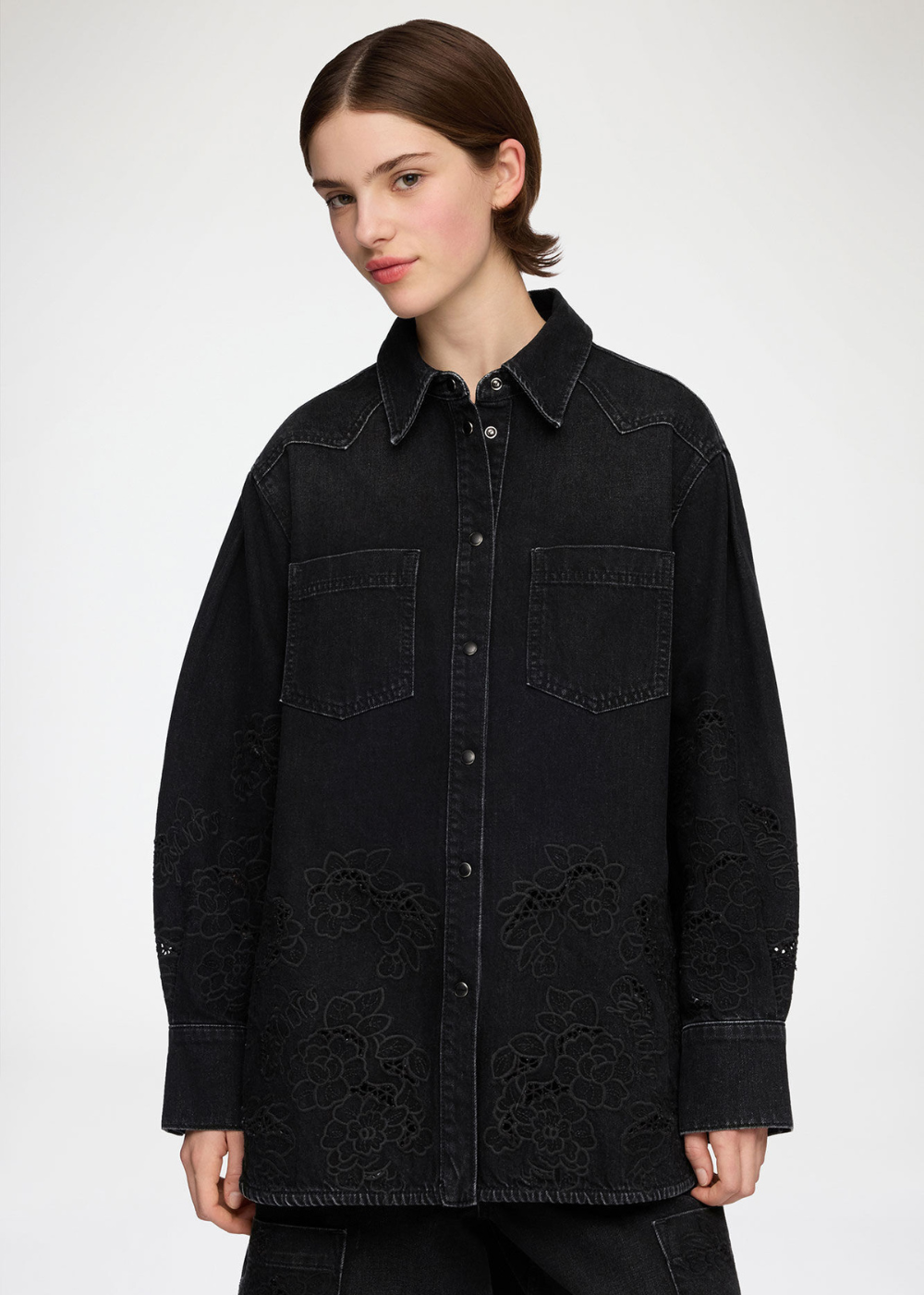 A model wearing a black denim button-up shirt jacket with tonal floral embroidery on the front, photographed against a light gray studio background.