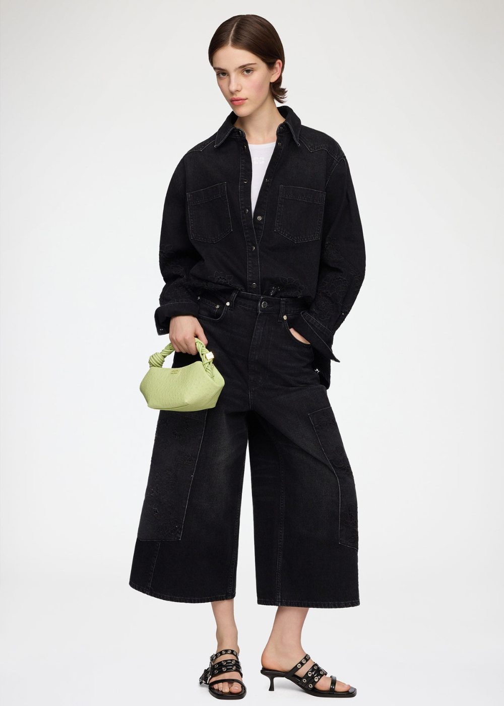 A model stands against a light gray studio background wearing a black denim-style button-up shirt, matching wide-leg cropped pants, a white top, black heeled sandals, and holding a small light green handbag.
