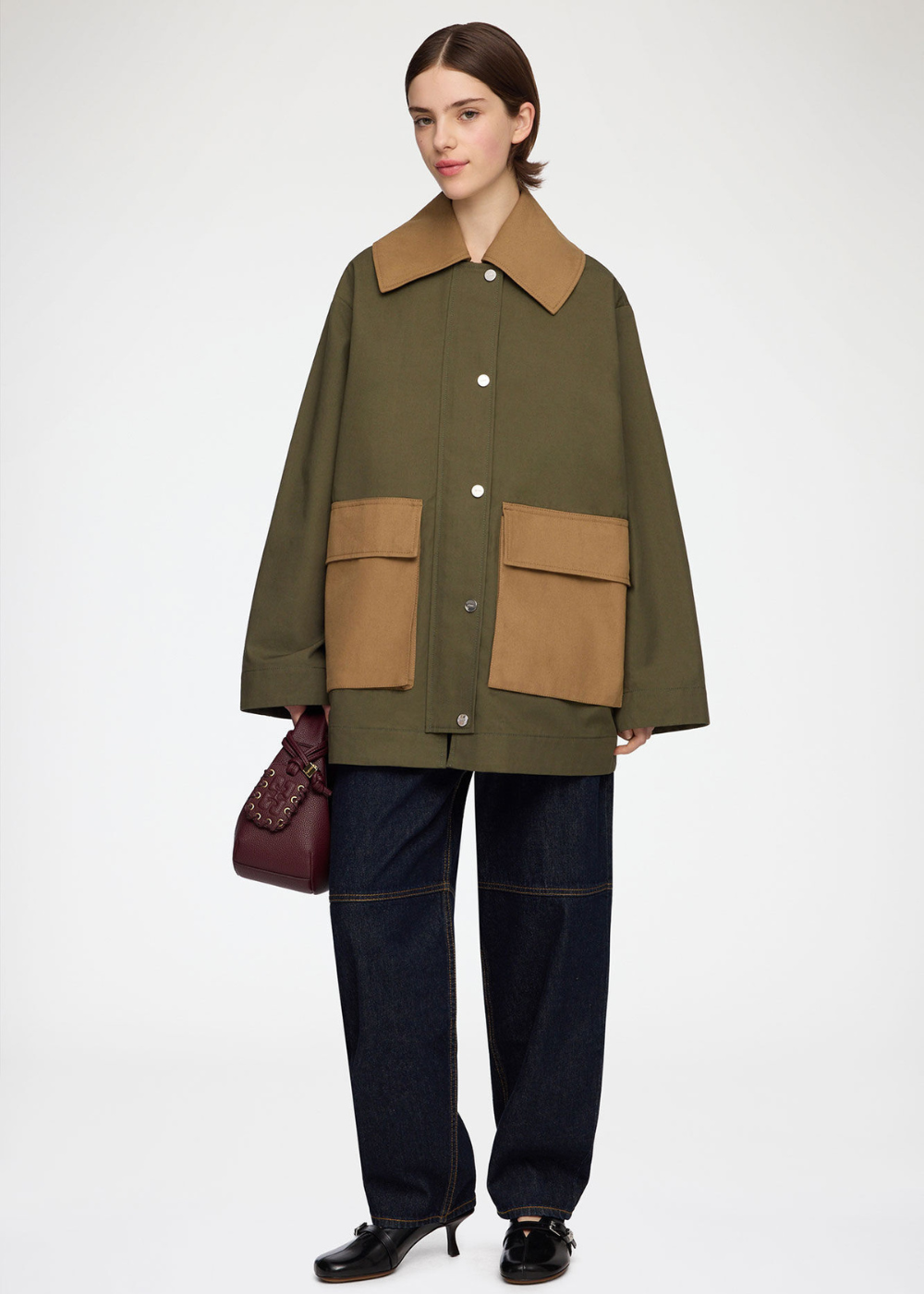 A model stands against a light gray studio background wearing an oversized olive green utility jacket with contrasting tan collar and front patch pockets, paired with dark blue jeans and black shoes, while holding a burgundy handbag.