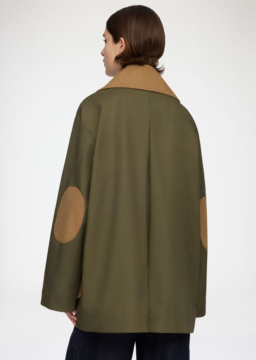 A woman shown from the back wearing an olive green utility-style jacket with tan circular elbow patches and a tan collar, against a plain light gray studio background.
