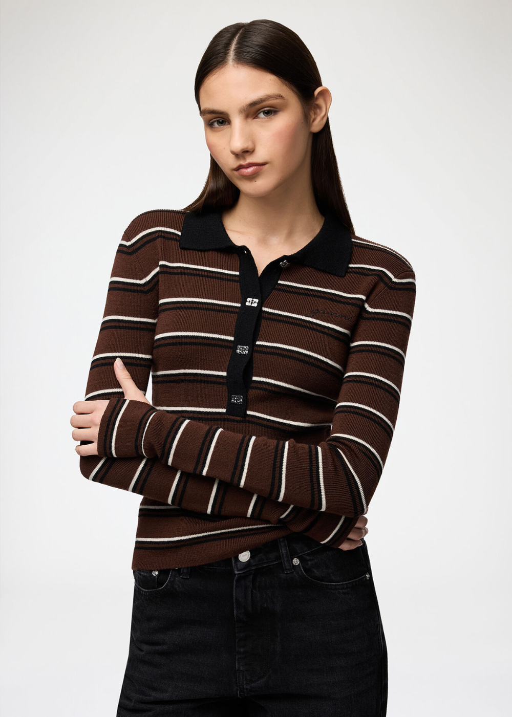 A woman modeling a brown long-sleeve knit polo shirt with thin white and beige horizontal stripes, a black collar, and a black button placket, paired with black jeans against a light gray studio background.