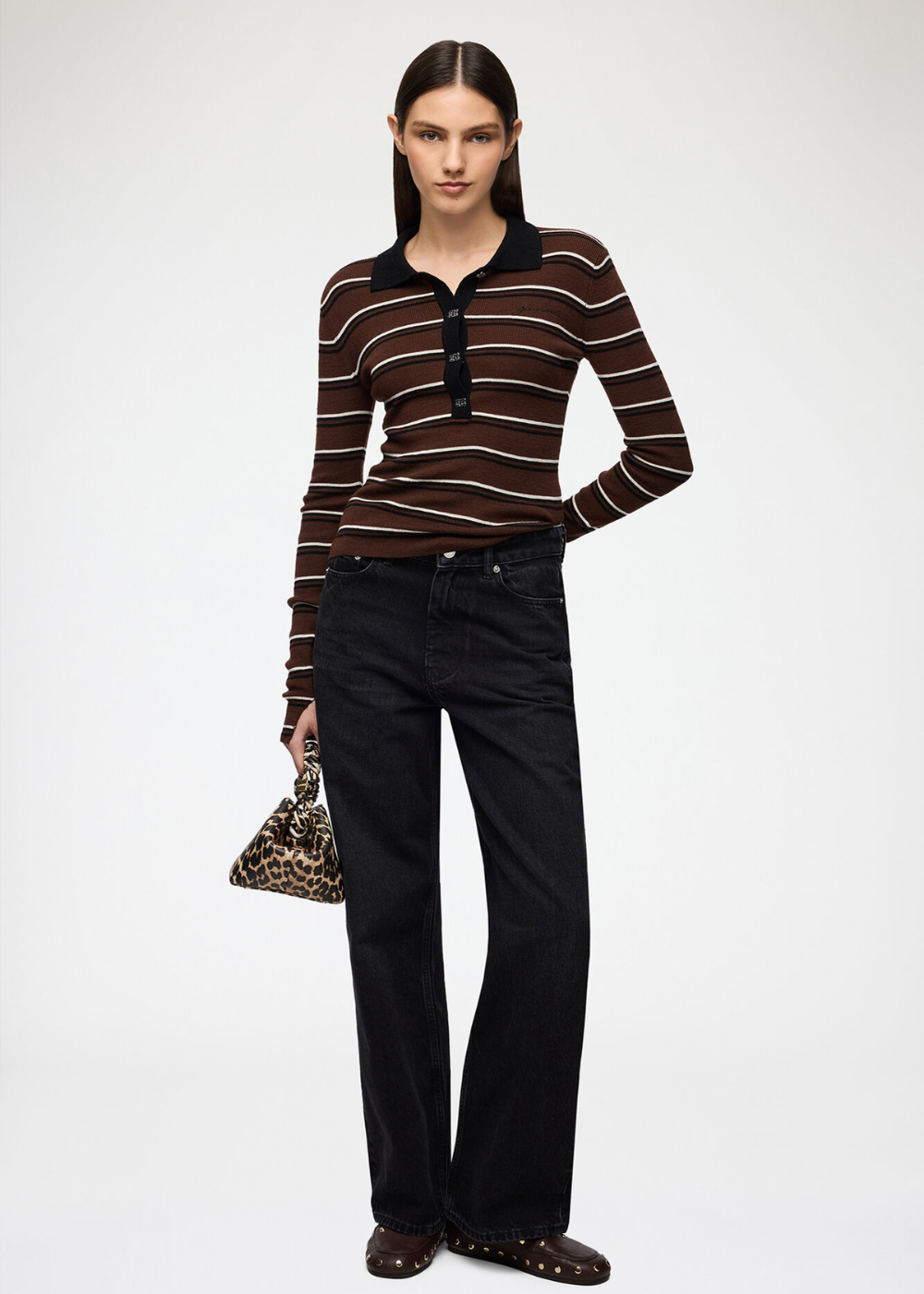 A model stands against a light gray studio background wearing a fitted long-sleeve polo-style top with horizontal brown, rust, and cream stripes, paired with black wide-leg jeans and dark pointed flats. She holds a small leopard-print handbag.
