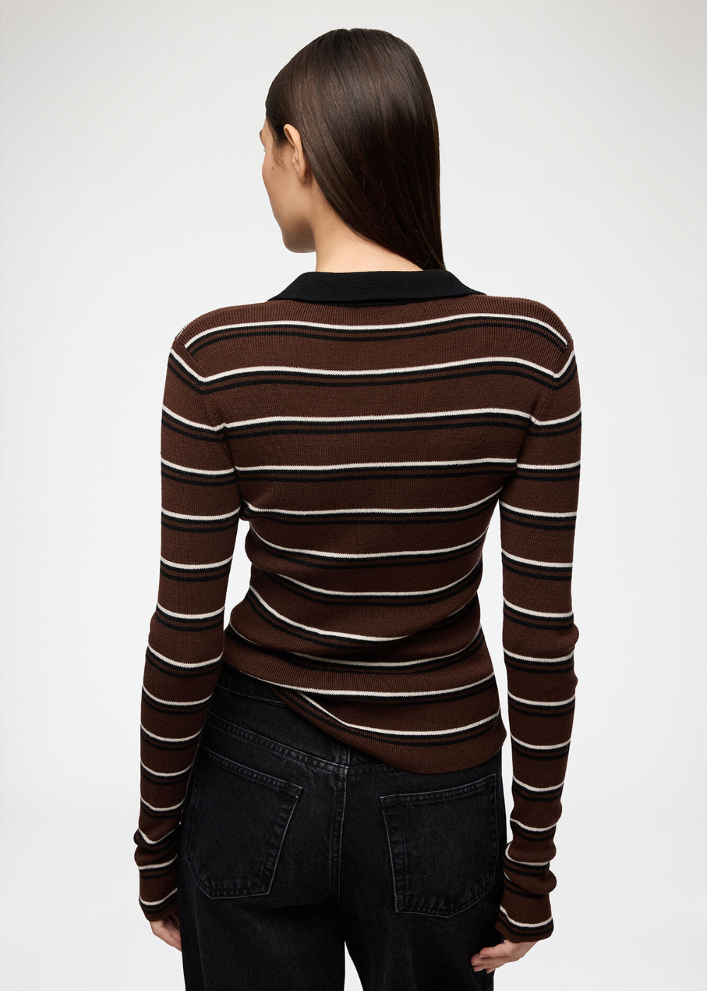 A woman shown from the back wearing a fitted long-sleeve knit top with horizontal brown, dark brown, and off-white stripes, paired with dark jeans, against a plain light gray studio background.