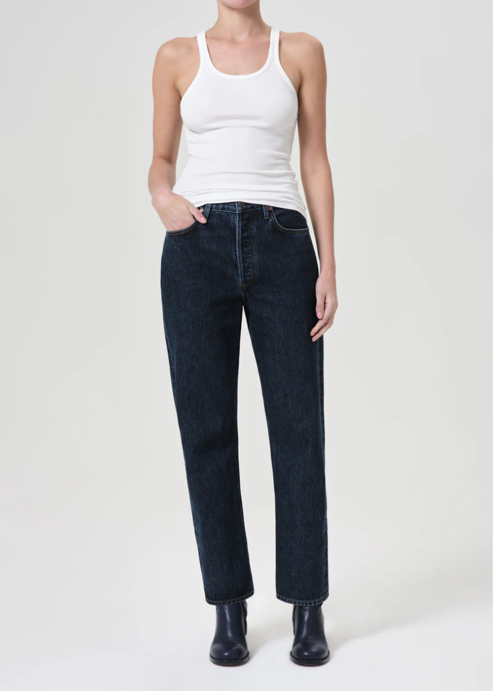 A model wearing dark wash straight-leg jeans, a white ribbed tank top, and black boots, standing against a light gray studio background.