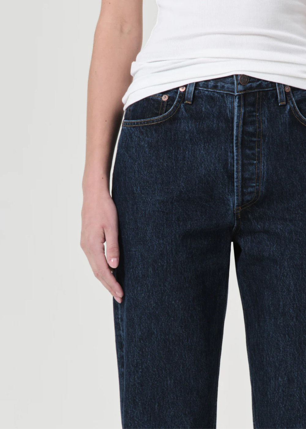 Close-up of a person wearing dark blue denim jeans and a white top against a light gray studio background.