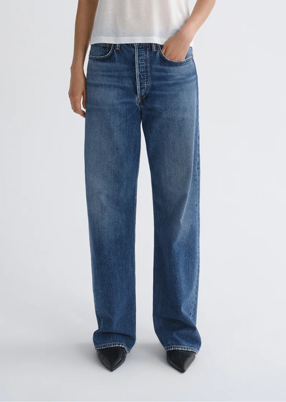 A person wearing blue wide-leg jeans with a white top and black pointed shoes, standing against a light gray studio background.