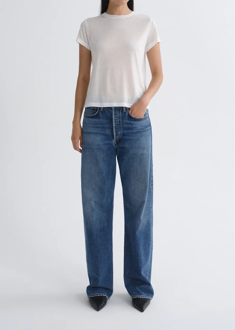 A model wearing a white short-sleeve crewneck t-shirt and high-waisted, wide-leg blue denim jeans, paired with black pointed-toe heels, against a light gray studio background.