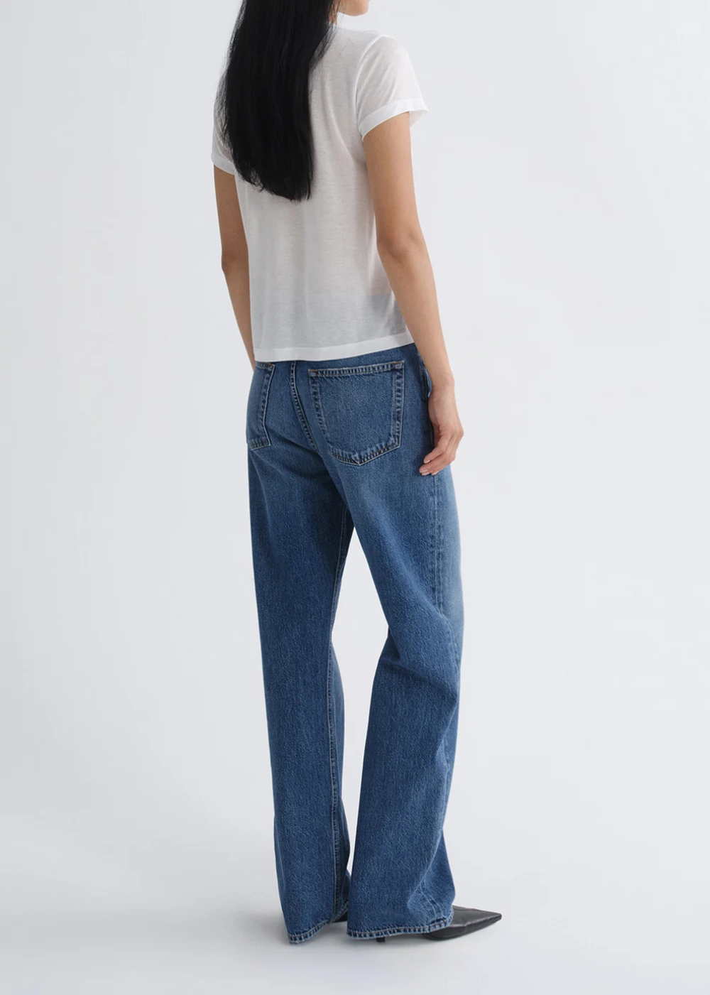 A model shown from the back wearing blue wide-leg jeans, a white short-sleeve t-shirt, and black shoes against a light gray studio background.