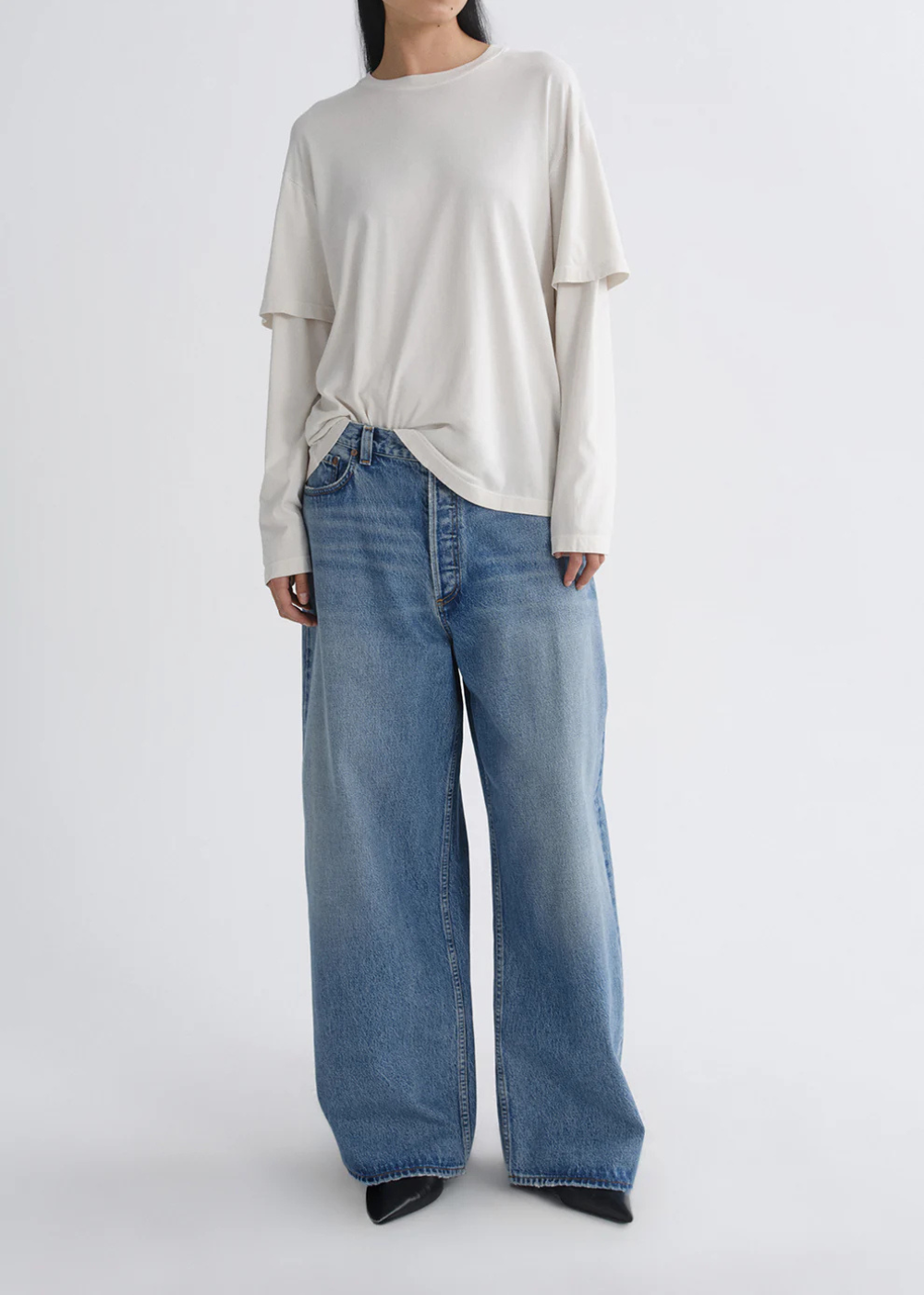 A model wearing a cream-colored, long-sleeve layered t-shirt and wide-leg blue denim jeans, standing against a plain light gray studio background.