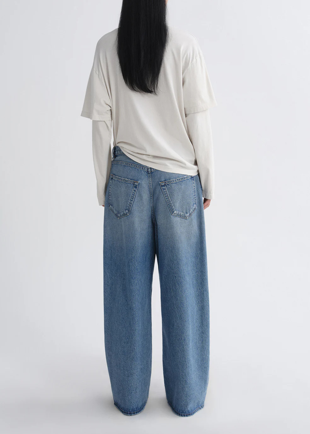 A person shown from the back wearing a cream-colored long-sleeve top and wide-leg blue denim jeans against a plain light gray studio background.
