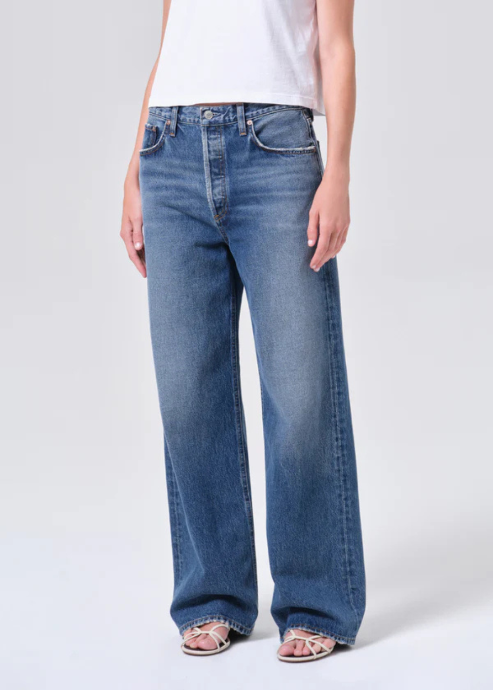 A model wearing a pair of wide-leg blue denim jeans with a faded wash, paired with a white sleeveless top and pink sandals, shown from the waist down against a light gray studio background.