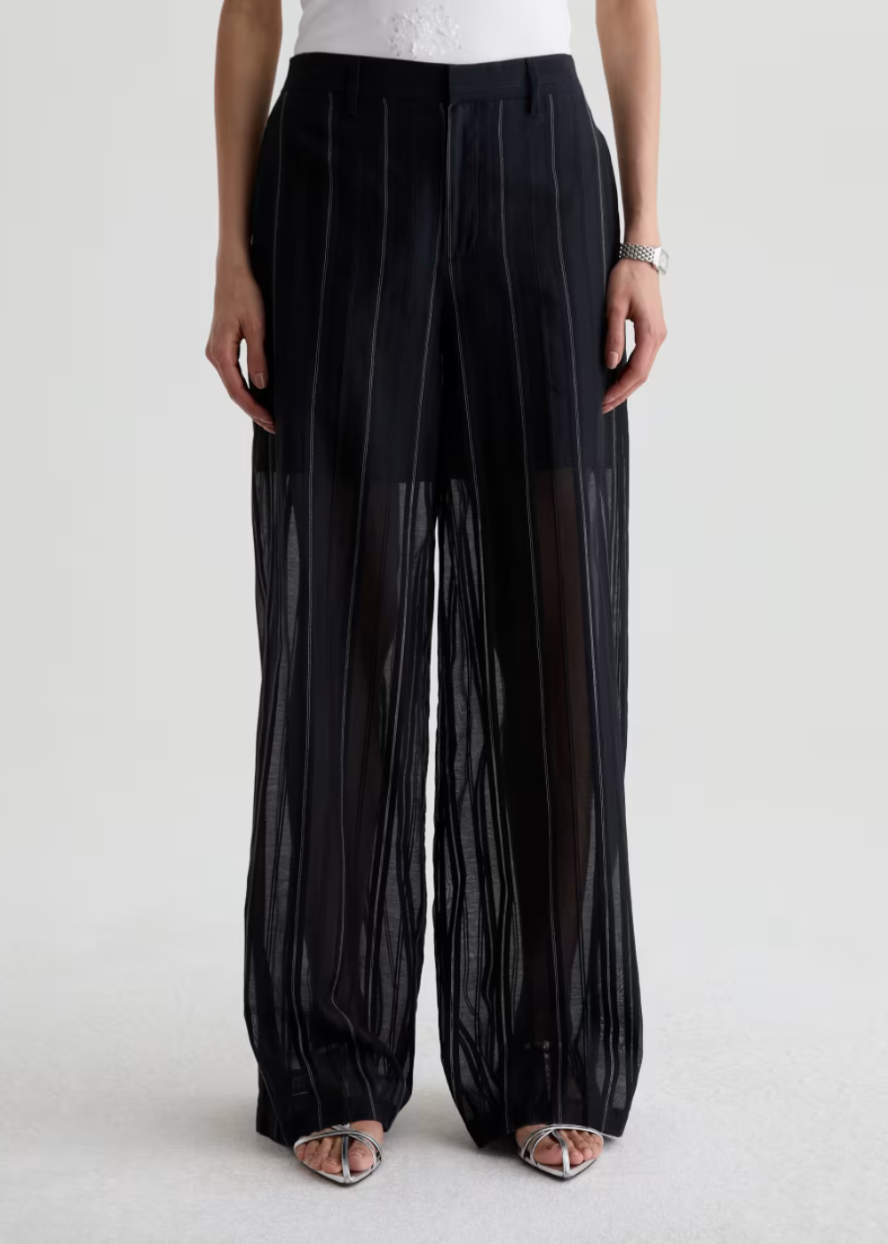 A model is shown from the waist down wearing black wide-leg pleated trousers made of sheer mesh over a solid black base, paired with a white top and metallic sandals.