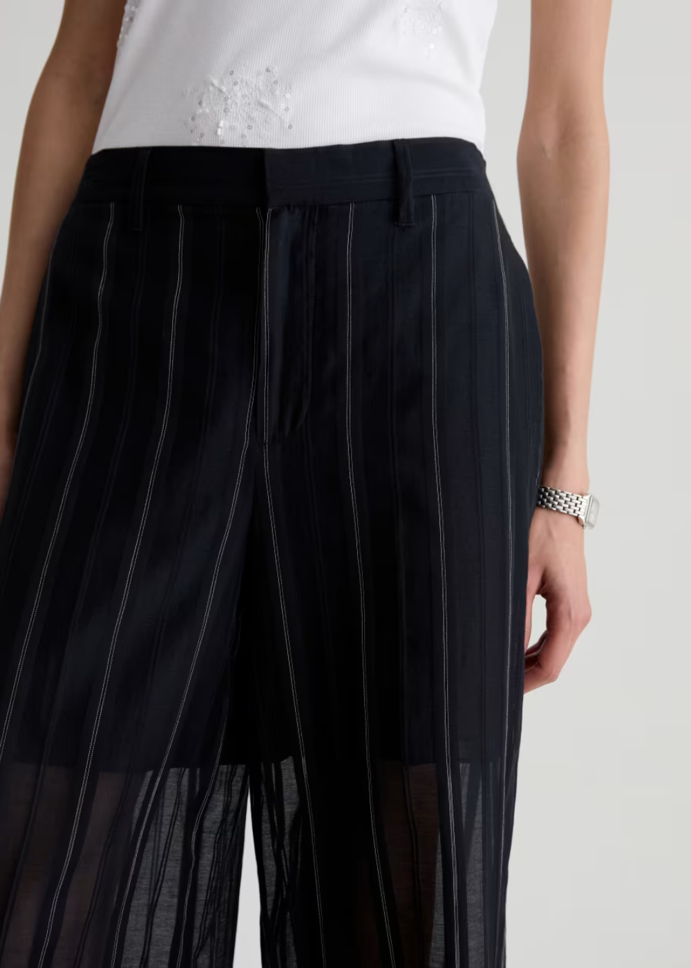 Close-up of a model wearing black wide-leg trousers with thin vertical white pinstripes and a sheer mesh lower panel, paired with a white top.