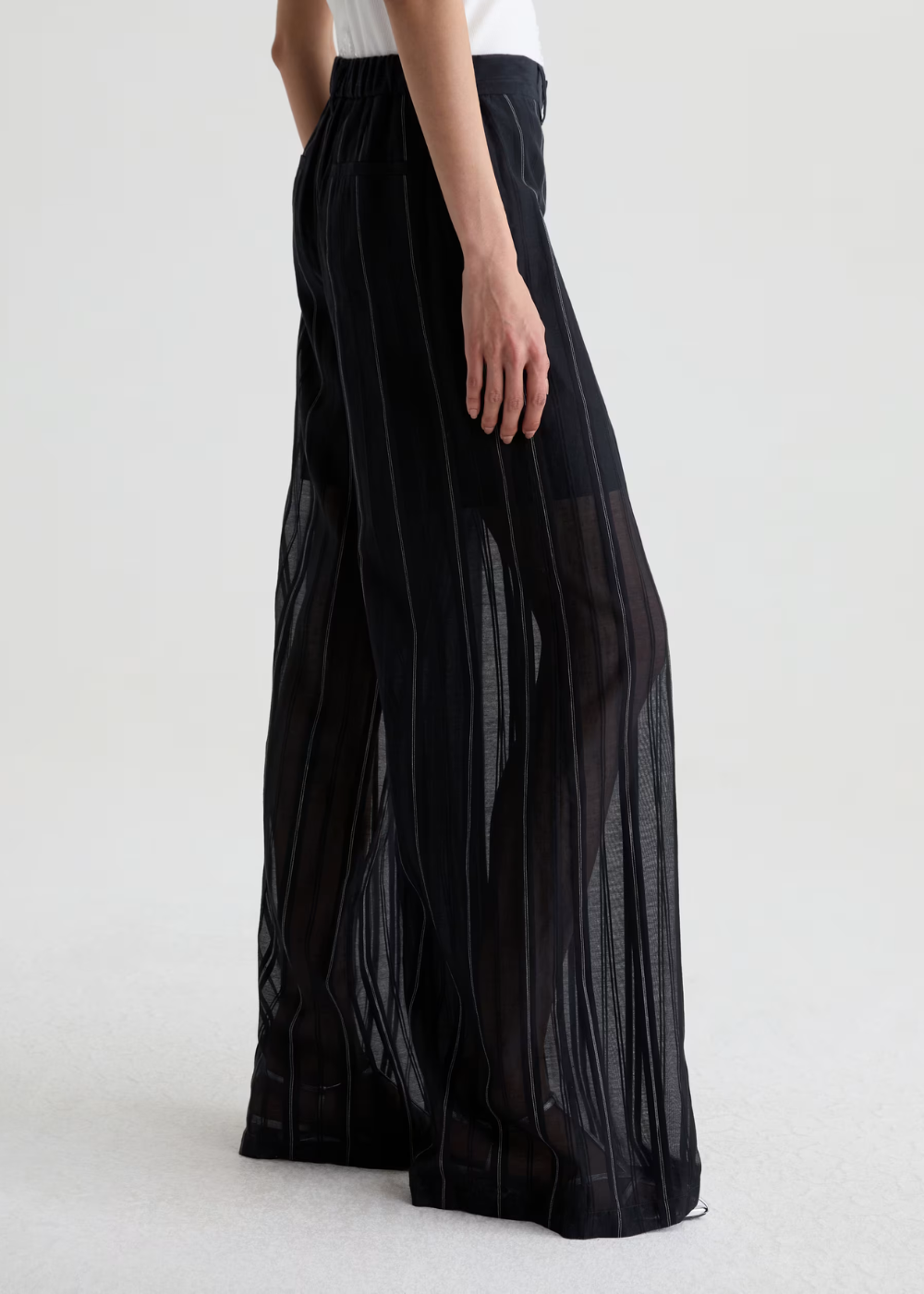 Black wide-leg pleated trousers with sheer mesh side panels, shown on a model from the side against a light gray studio background.