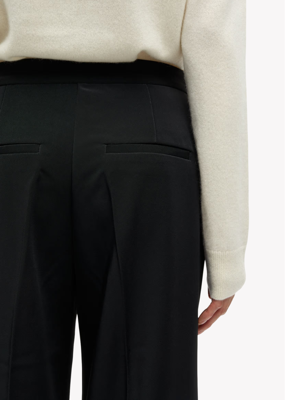 Close-up of a person wearing black wide-leg trousers with a black waistband and a cream ribbed sweater, shown from the back against a light gray studio background.