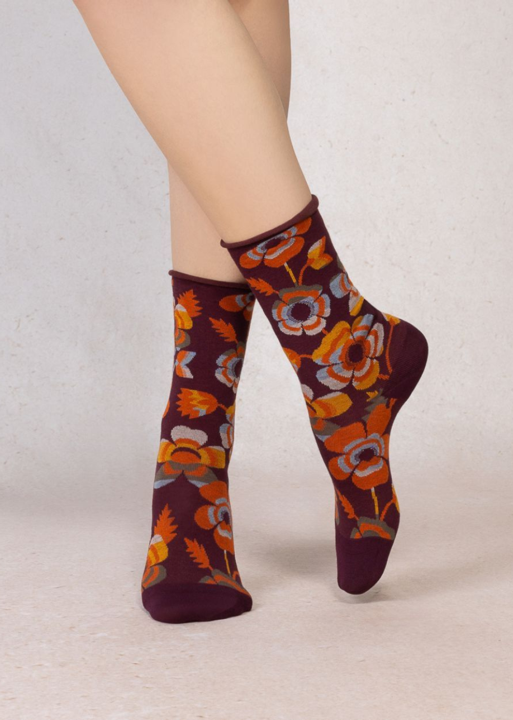 A pair of crew socks with a dark burgundy base and a vibrant orange, yellow, and gray floral pattern, shown on a model's legs against a neutral light beige background.
