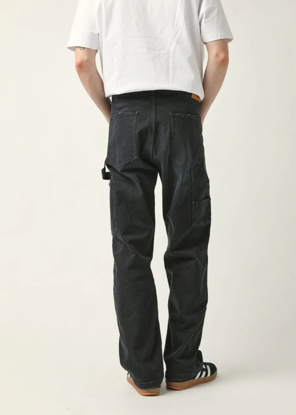 Rear view of a person wearing dark charcoal cargo pants, a white t-shirt, and black-and-white sneakers against a plain light background.
