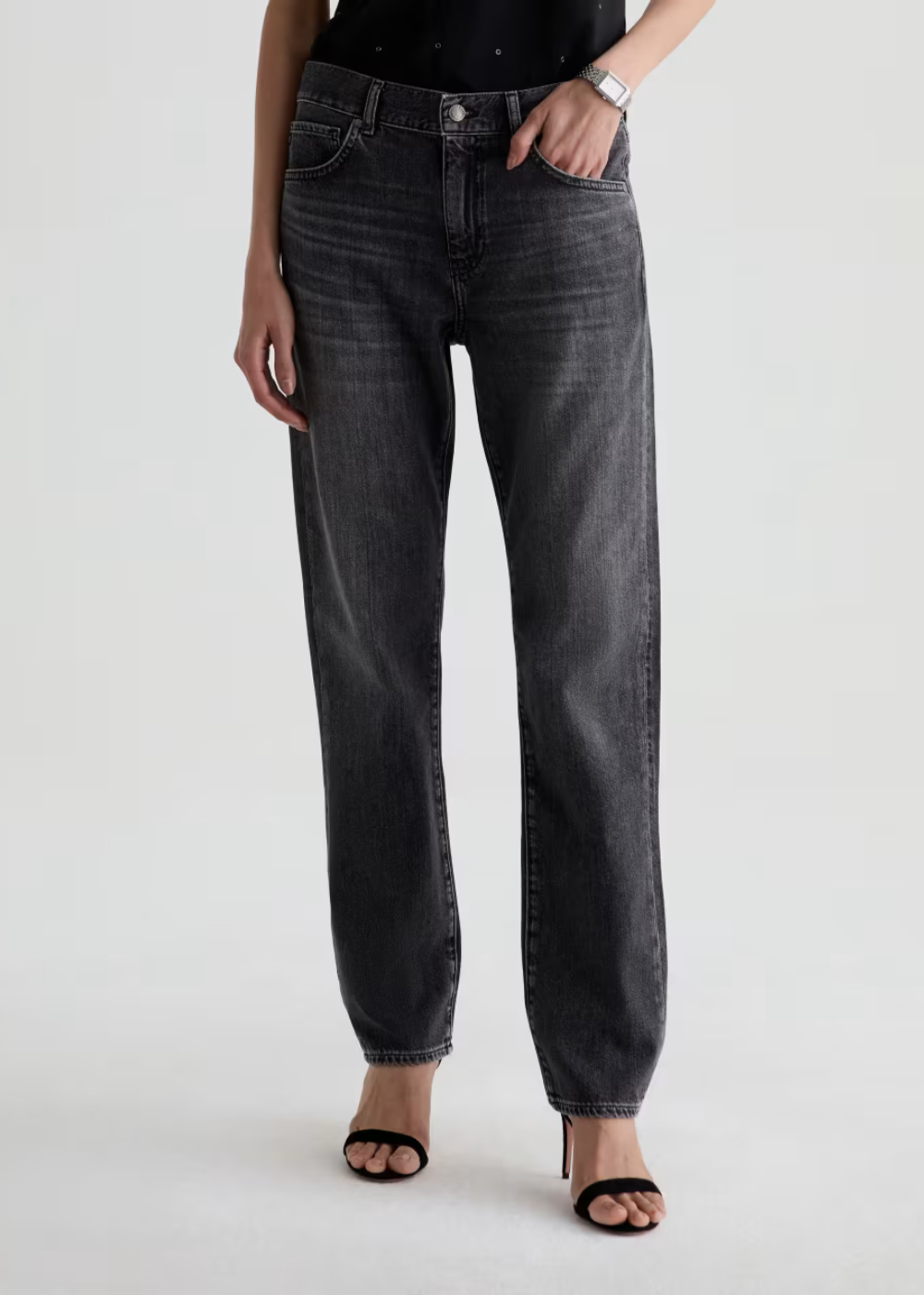 A model wearing a pair of dark gray washed straight-leg jeans, styled with a black top and black heeled sandals against a light gray studio background.