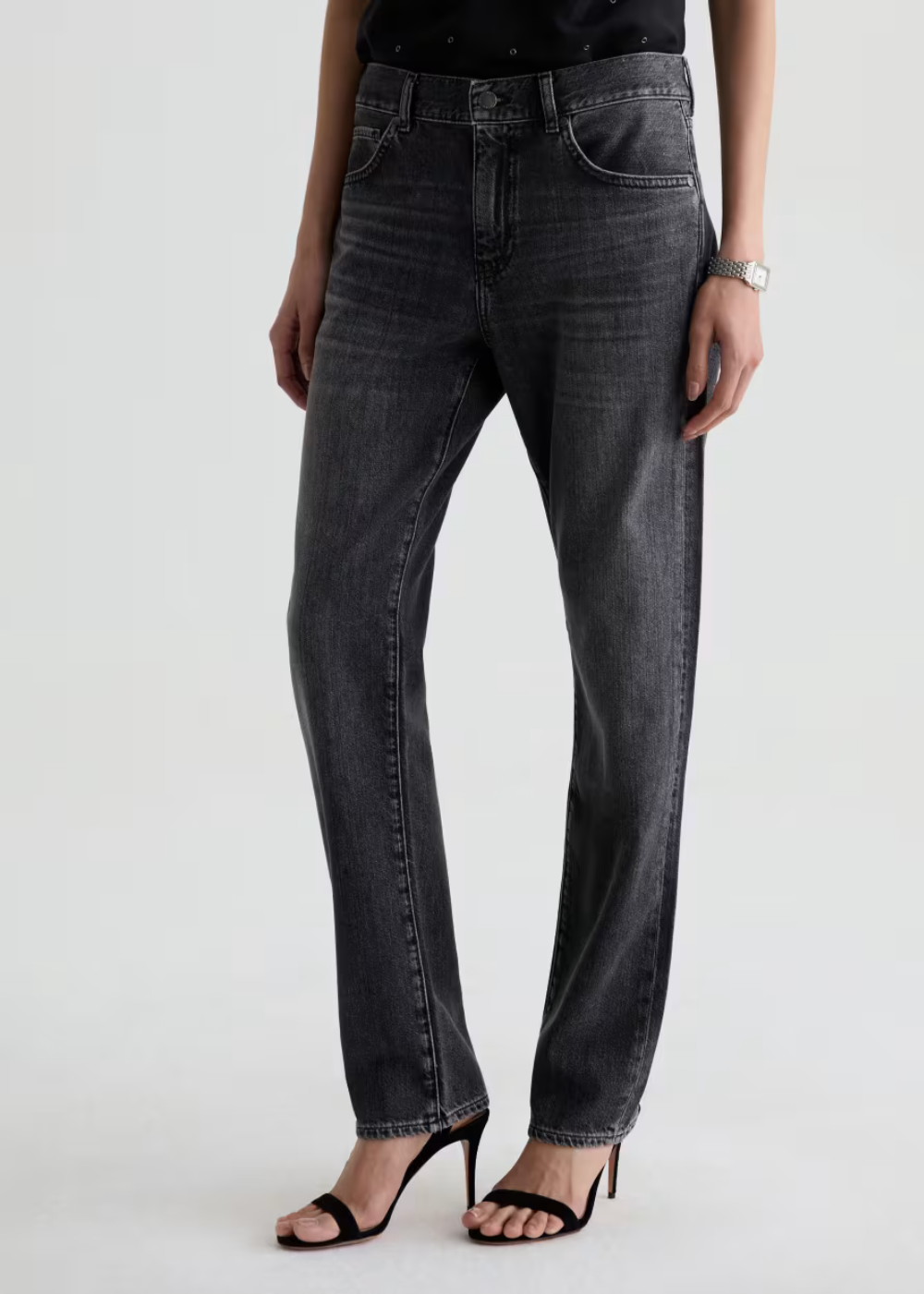 A model wearing a pair of dark gray washed straight-leg jeans with black heeled sandals, shown from the waist down against a light gray studio background.