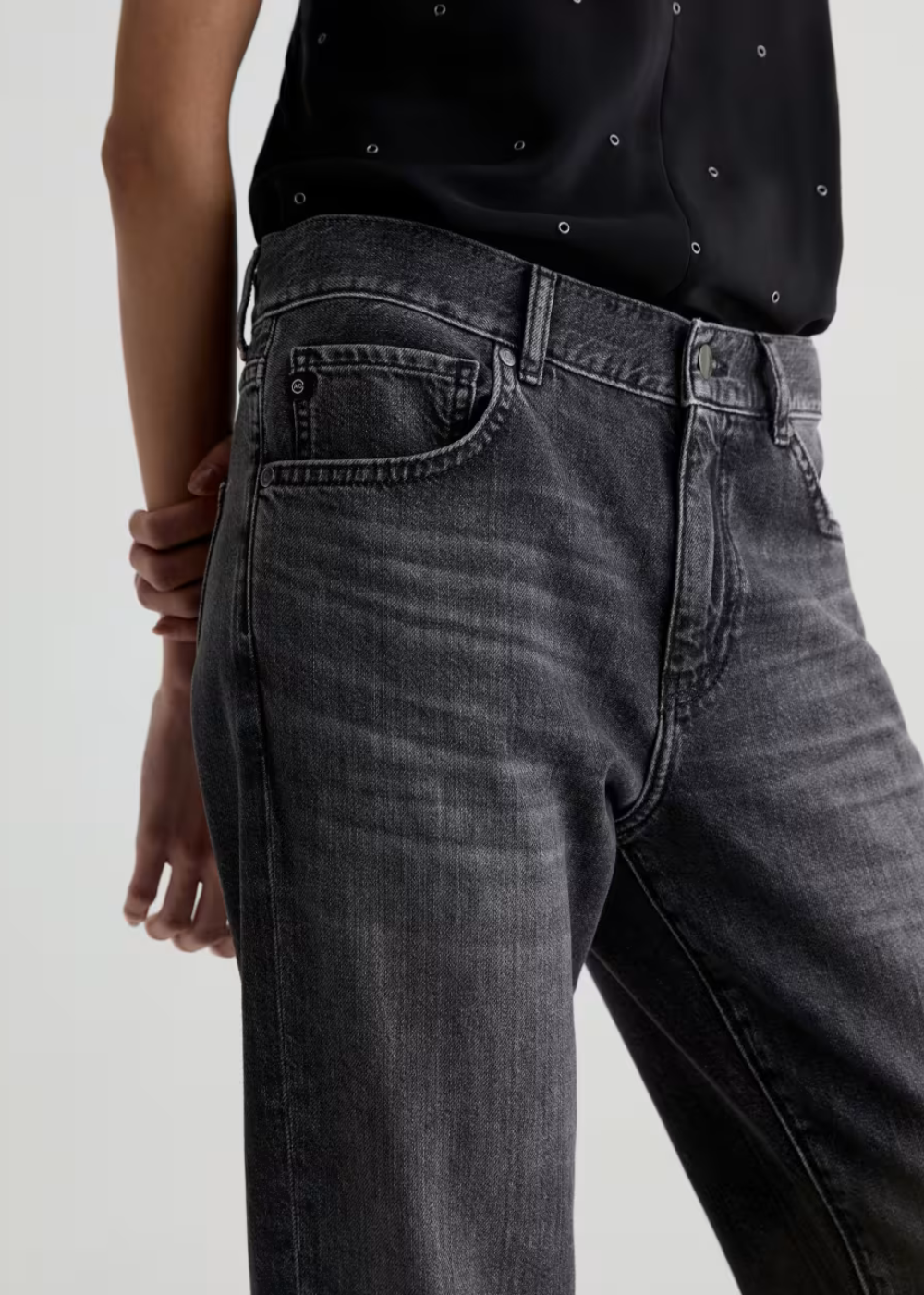 Close-up of a person wearing dark gray washed denim jeans and a black top with small silver embellishments, shown against a light gray studio background.