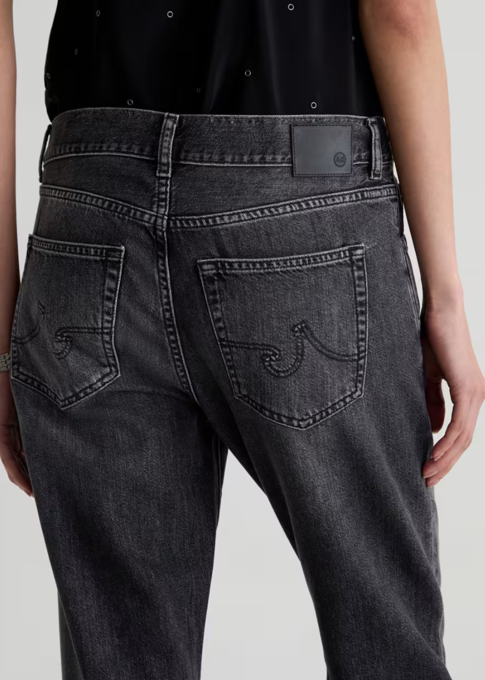 Close-up back view of a person wearing dark gray washed denim jeans with decorative stitching on the back pockets and a black top.