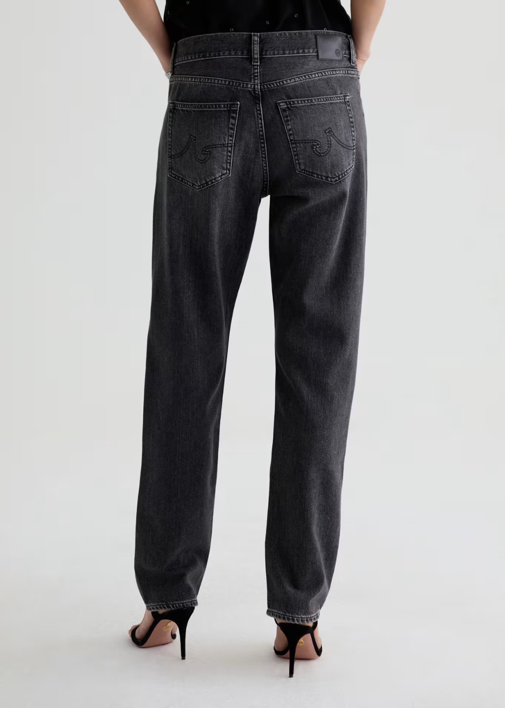 Rear view of a person wearing dark gray washed denim jeans with embroidered back pocket details, paired with black high-heeled sandals against a light gray studio background.