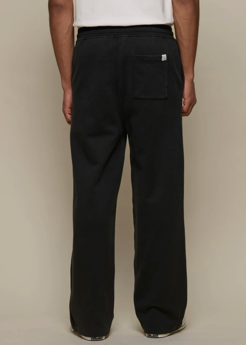 A person shown from the back wearing loose black wide-leg pants with a back pocket, against a plain beige studio background.