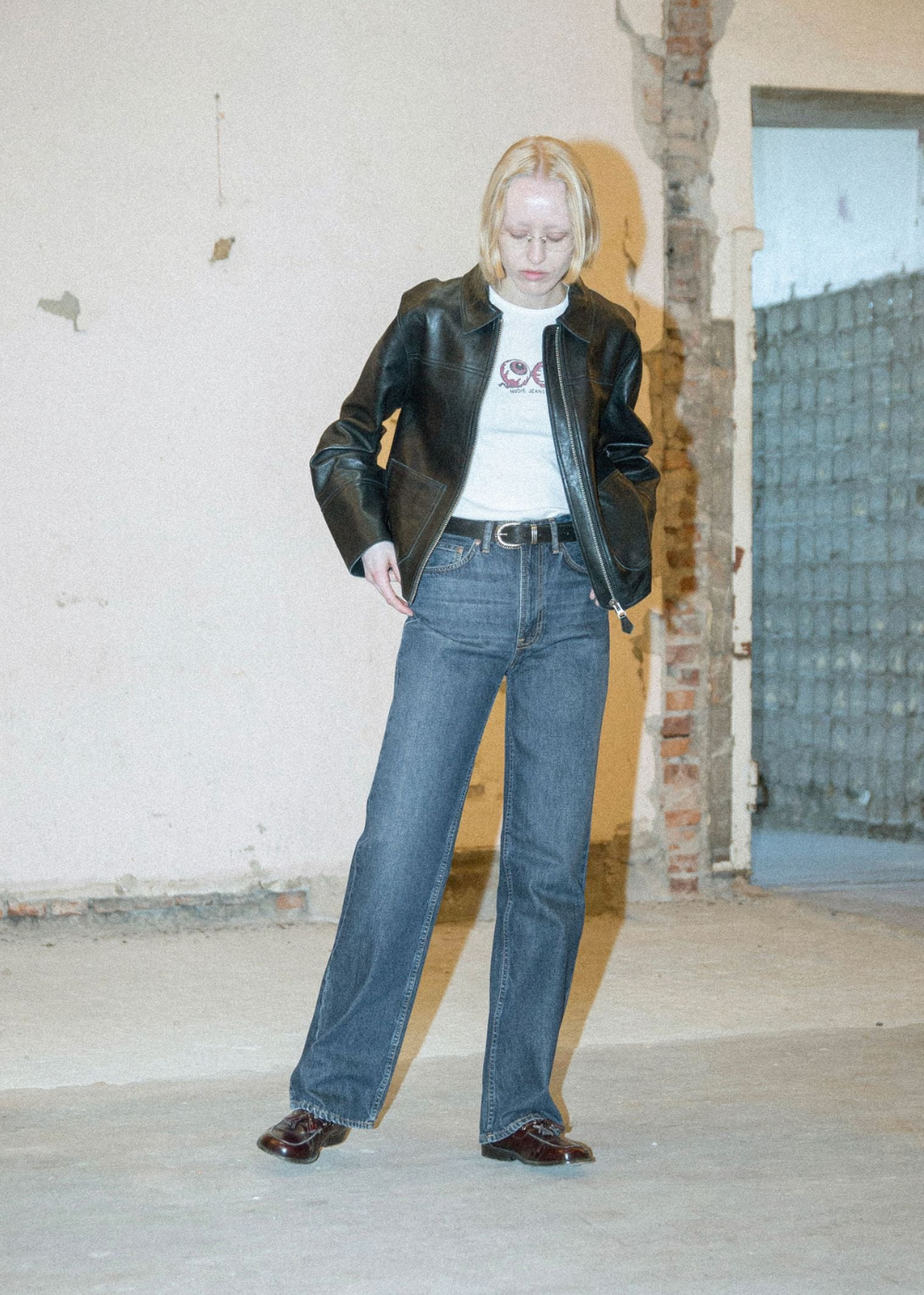 A model stands in a worn industrial room wearing a black leather jacket, a white t-shirt with a graphic, wide-leg blue jeans, and brown leather shoes.